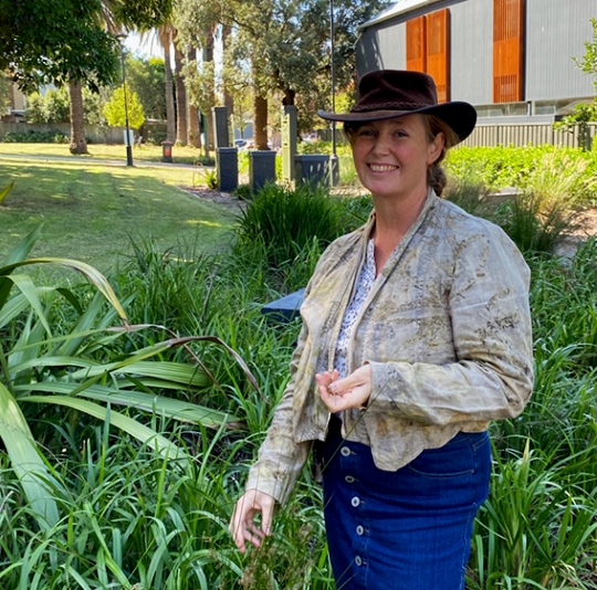 Smiling person in a wide-brimmed hat standing in front of dense low plants in an urban setting