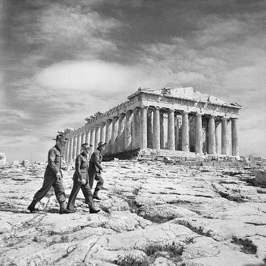 Black and white historical image of soldiers walking on gravel in front of the acropolis