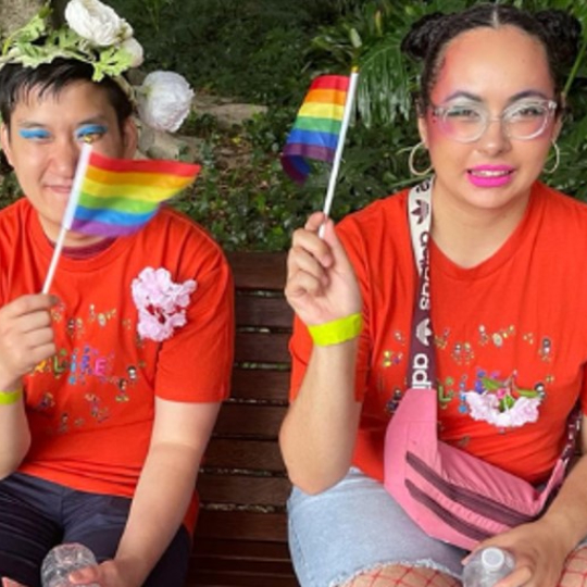 Two people sitting on a bench waving rainbow flags. One person has white flowers in their hair and blue eyeshaow and one is wearing bright red lipstick