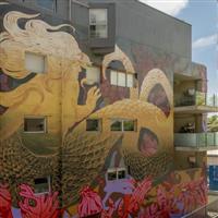 An apartment block with a large gold mermaid mural painted across the entire facade, against a background of pink and purple flannel flowers