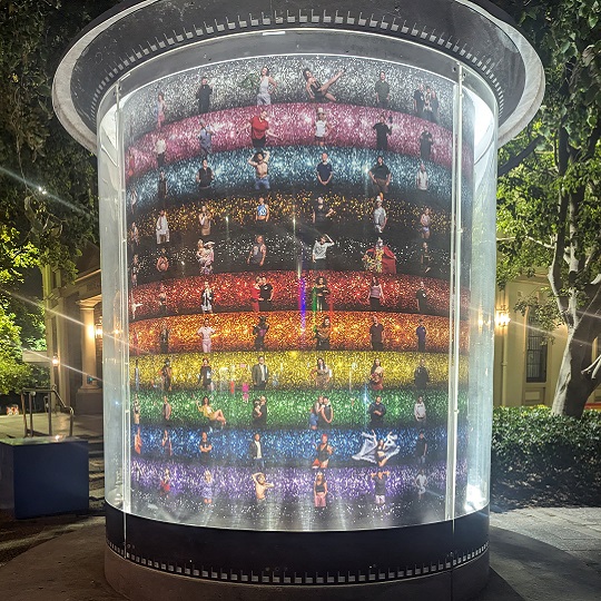 An image of Newtown ArtSeat, a cylindrical public art installation with a transparent outer shell, with concrete seating at the base. Showcasing a vibrant rainbow-colored glitter background depicting the pride progress flag. Photographic images of people from the Newtown LGBTQIA+ community are arranged in dynamic poses across the surface. The image is taken at night and the artwork is illuminated by up lights.