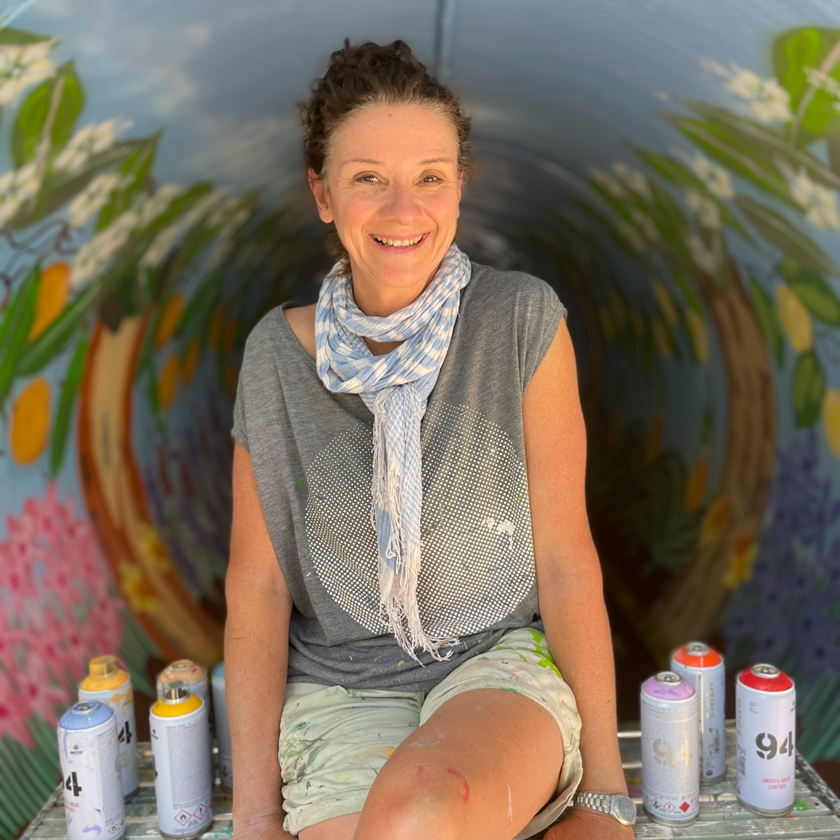 A headshot photograph of a woman sitting cross legged on a painting bench. She sits smiling in front of a painted tunnel. 