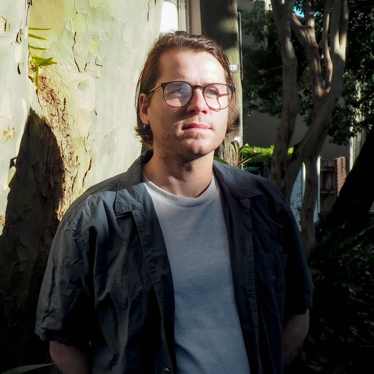 A headshot photograph of a man with shoulder length brown hair and glasses stands in front of a gum tree.