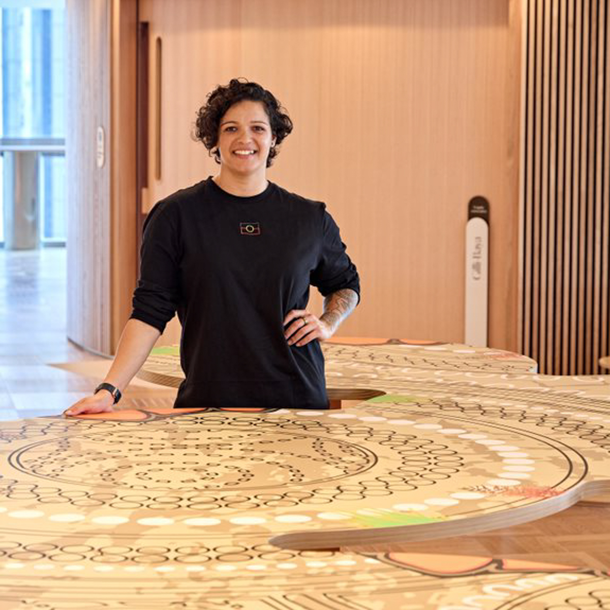 A smiling woman wearing a black shirt stands in a wooden room, in front of an artwork.