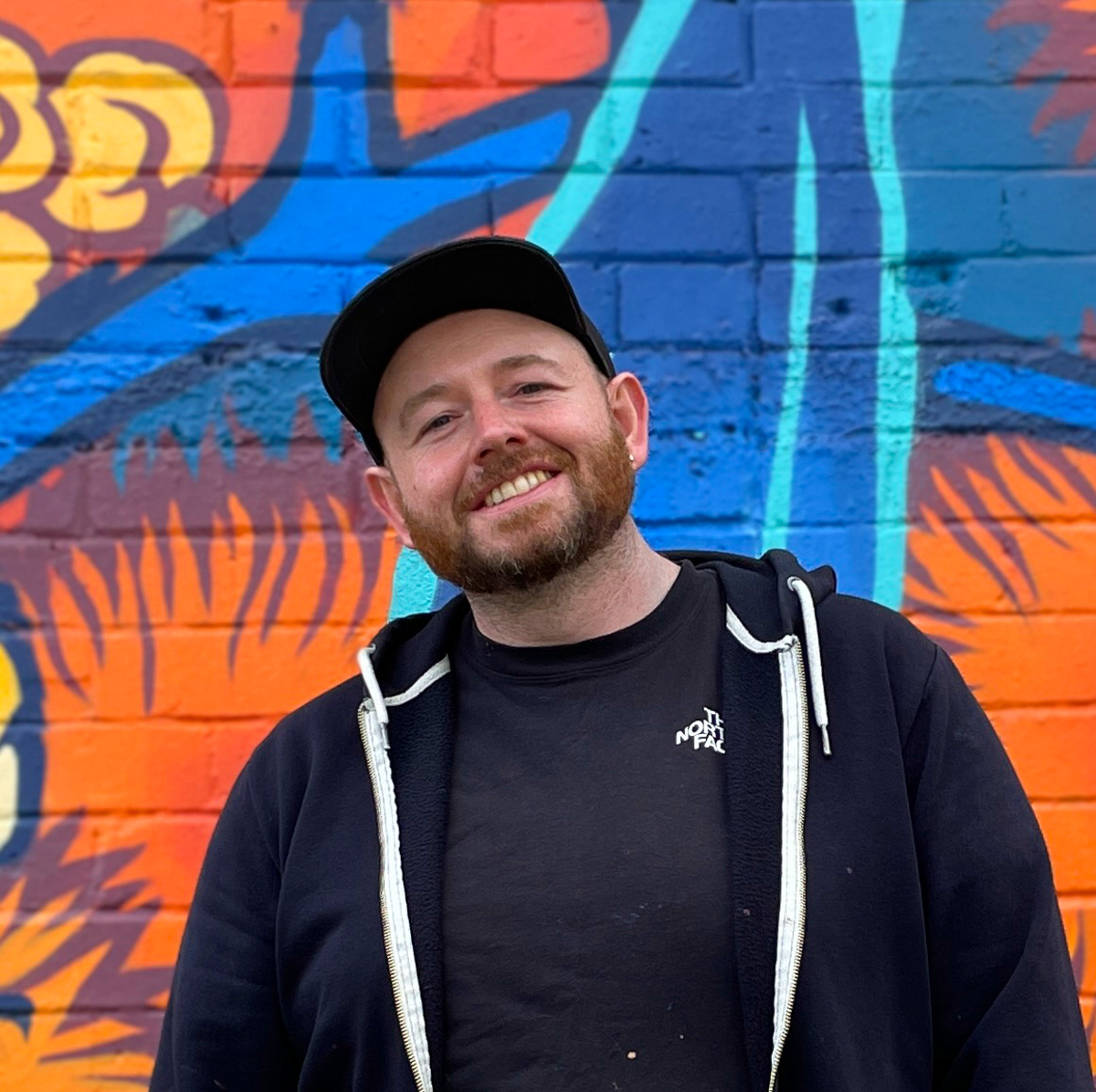 A headshot photograph of a smiling man standing in front of a colourful mural. He wears a cap.  