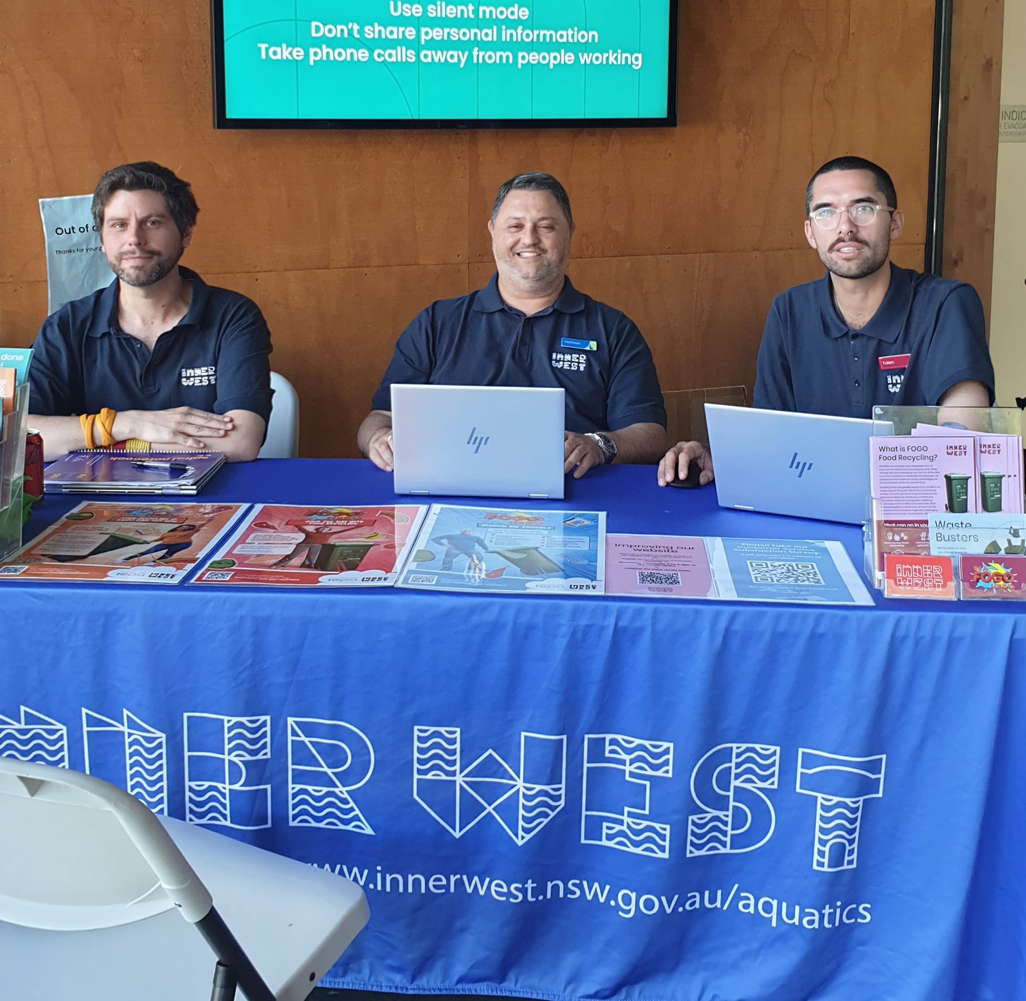 Three smiling customer service staff sitting at a tabled covered in a bright blue tablecloth with an Inner West logo on it