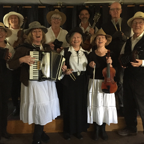 A collective of people in brown and white holding various instruments stand and sing happily together