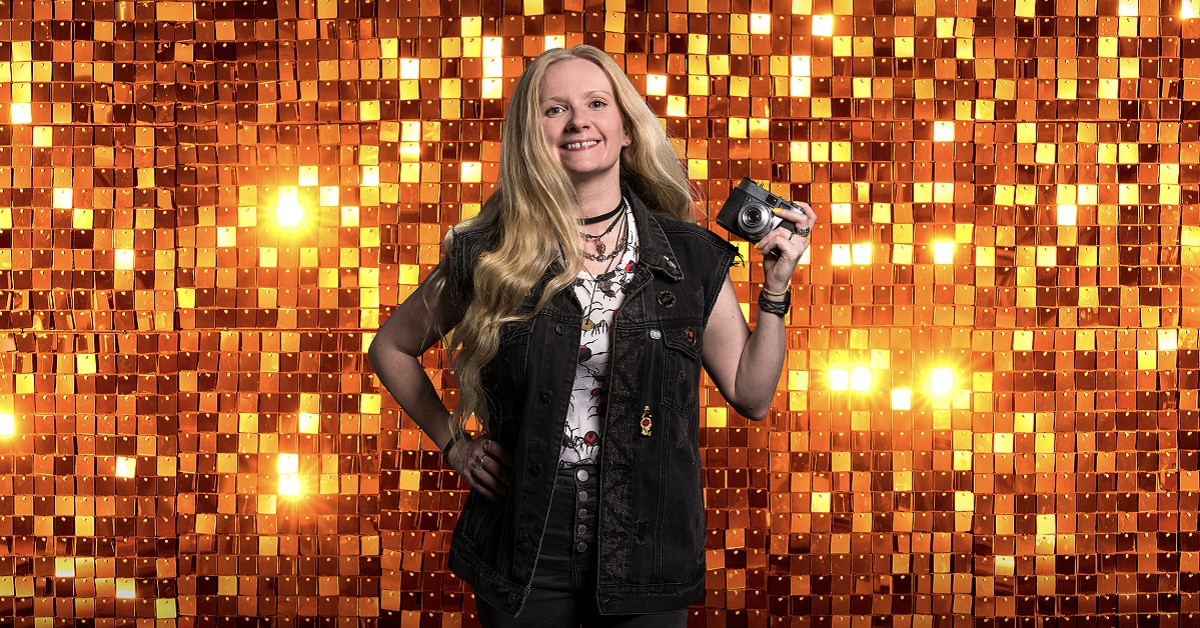 Image of Rhiannon Hopley smiling and holding a camera up in left hand in front of a gold sequined tiled background glinting in the light, Rhiannon has long blond hair and is wearing a sleevless black denim jacket, black jeans and white vest with a black and red print