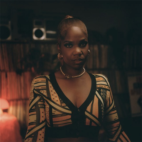 A woman in a dimly-lit room with a wall of vinyl records stored behind.