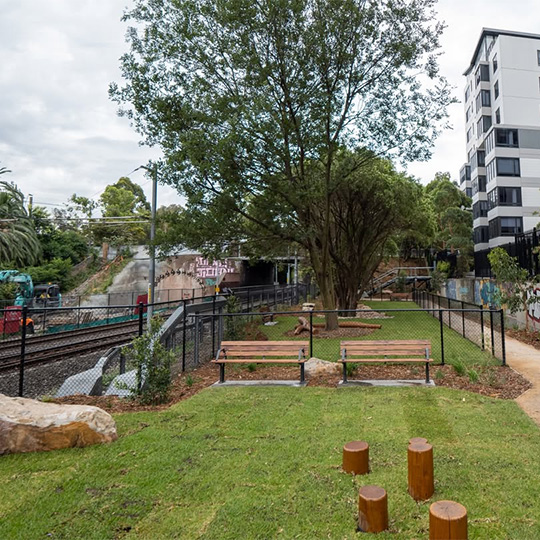 A fenced off-leash dog exercise area in a slender urban park, positioned between a light rail line and medium-rise buildings.