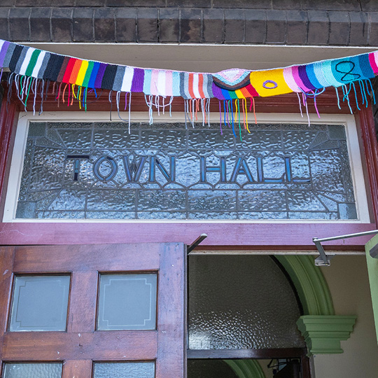 Knitted bunting of an array of community pride flags strung above an ornate wooden door with 'Town Hall' written in the stain glass panel.