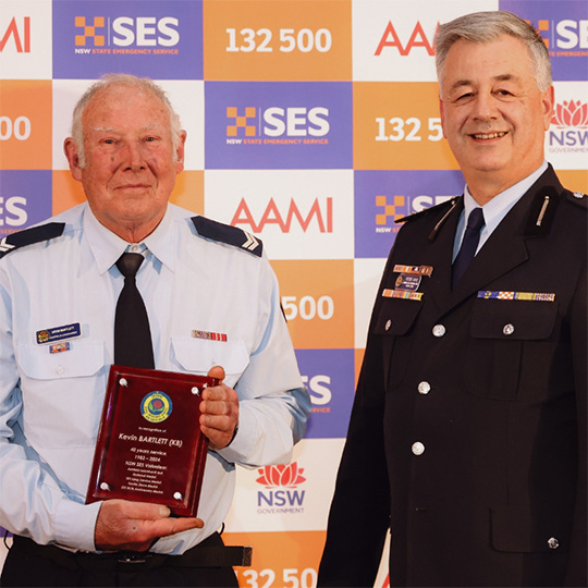 Two SES members in uniform stand against a media wall with SES logos on it. One holds an award