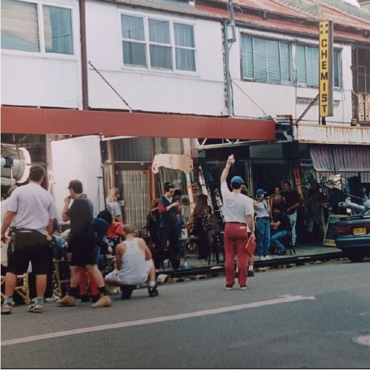 A bustling late-20th-century scene of people and a film crew in a small inner-suburban commercial area
