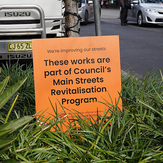 Orange sign nestled in green plants reads "These works are part of Council's Main Streets Revitalisation program"