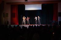 Four performers on stage in business attire set the scene for their next dance showcase - one reads a newspaper, one checks the time on their watch and the other two look towards each other