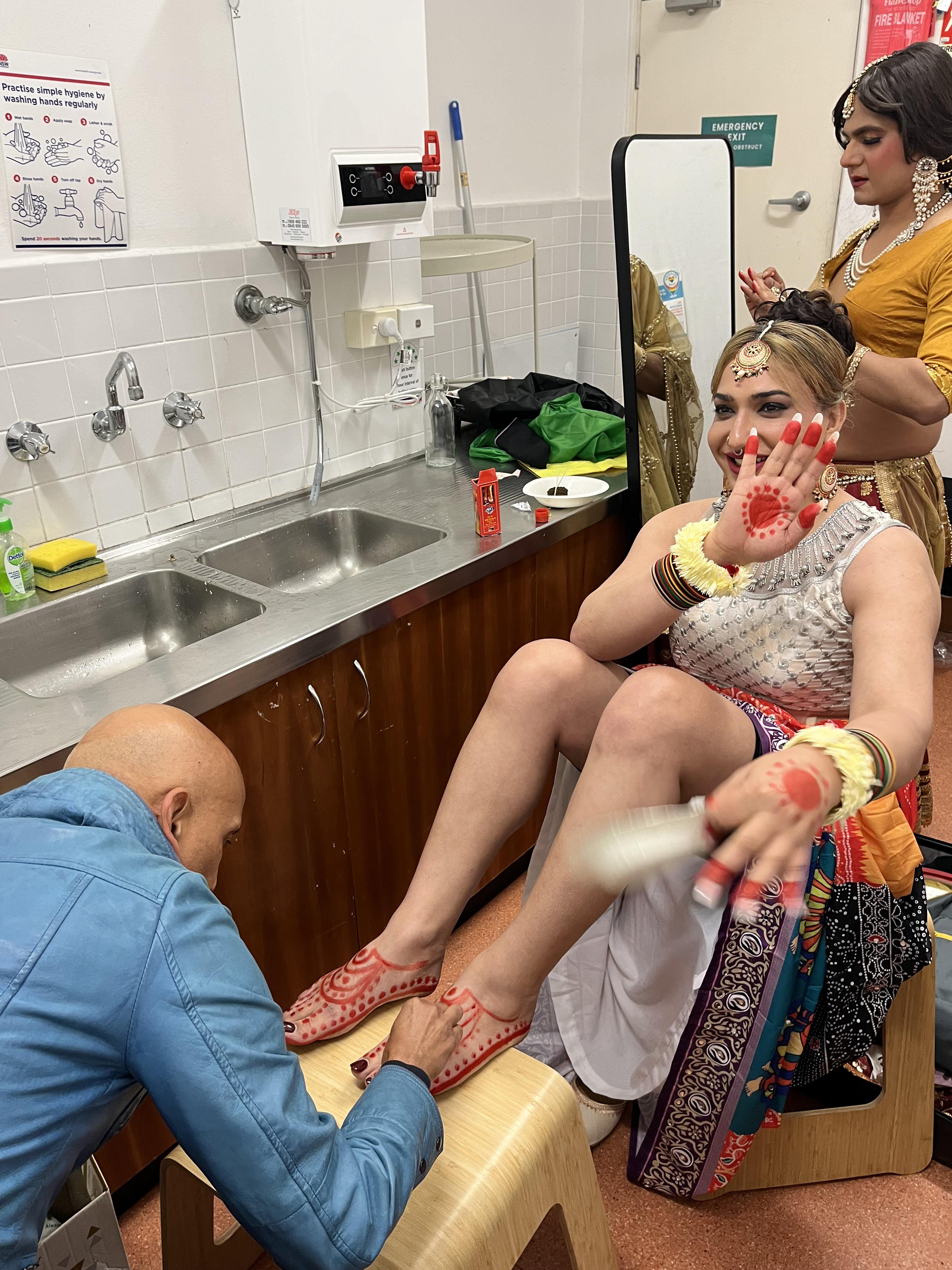 Photo of Satrang performer @sanakhantrans getting ready - they have red paint being applied to their hands and feet by another artist