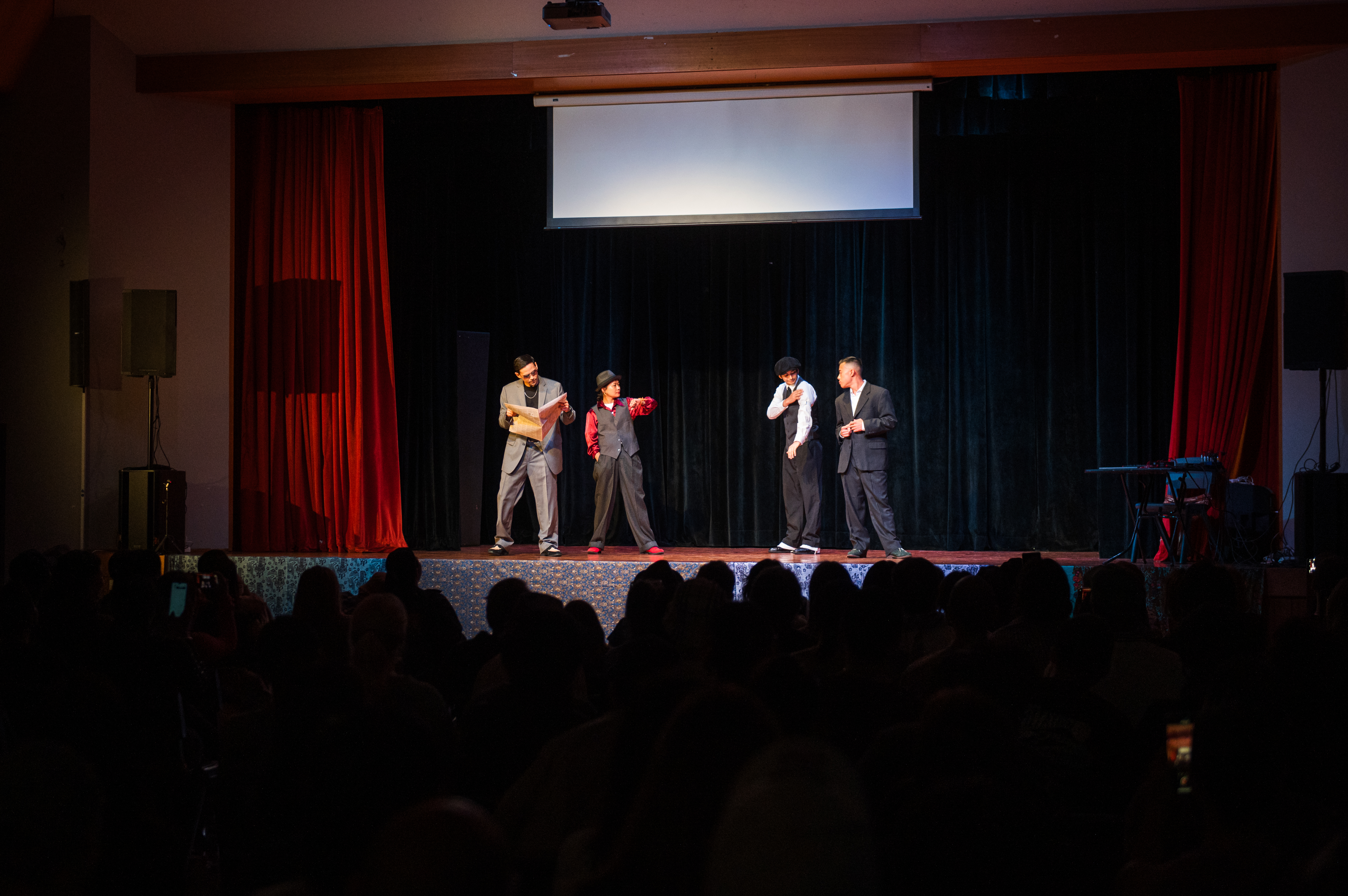 Photo of four dancers on stage wearing business attire, one read a newspaper, one checks the time on her wristwatch, the other two look to be talking to each other