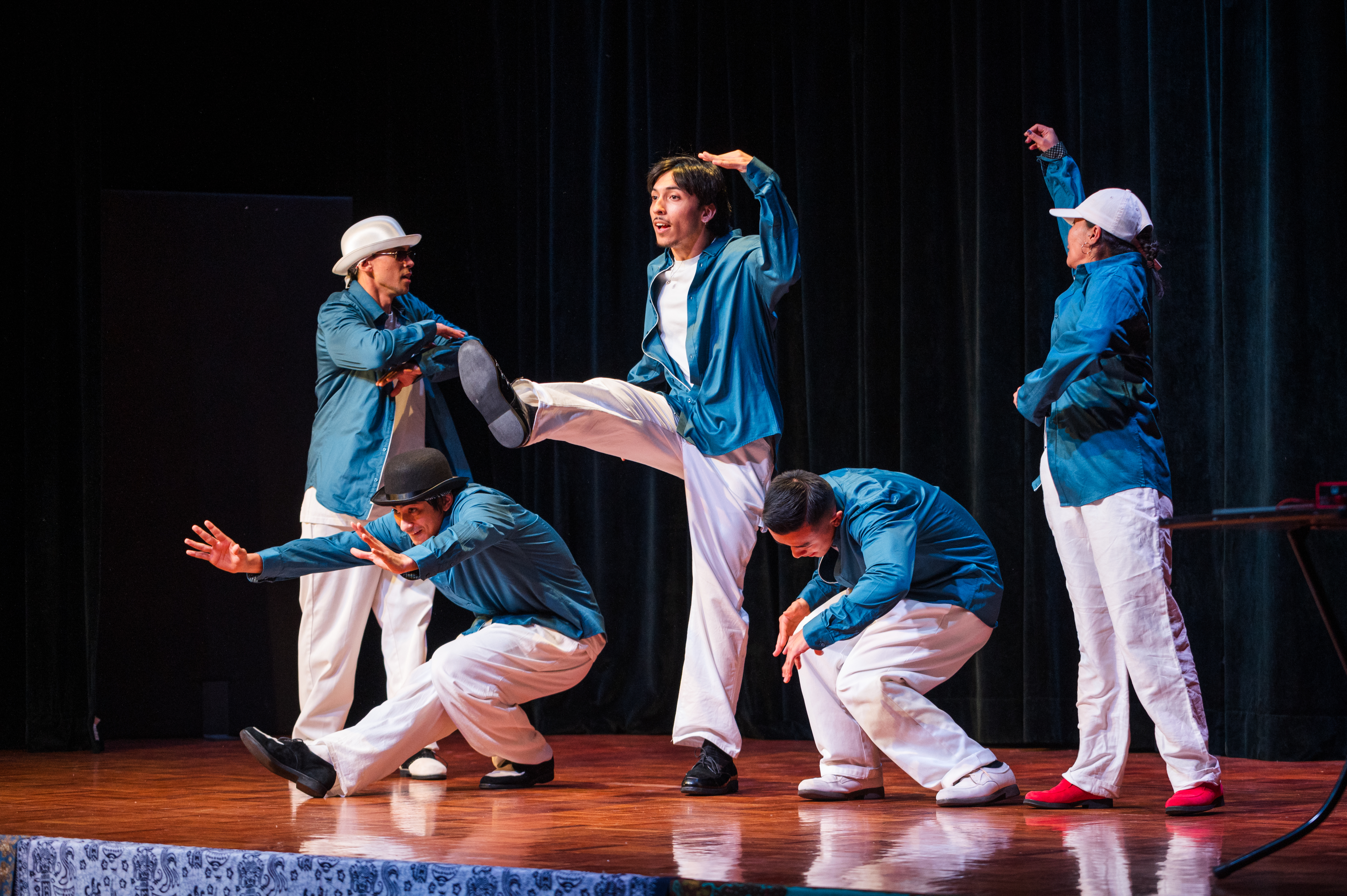 Photo of five Stale Biskits perfromers on stage wearing white pants and teal blue shirts, each is mid pose in popping style dance pose