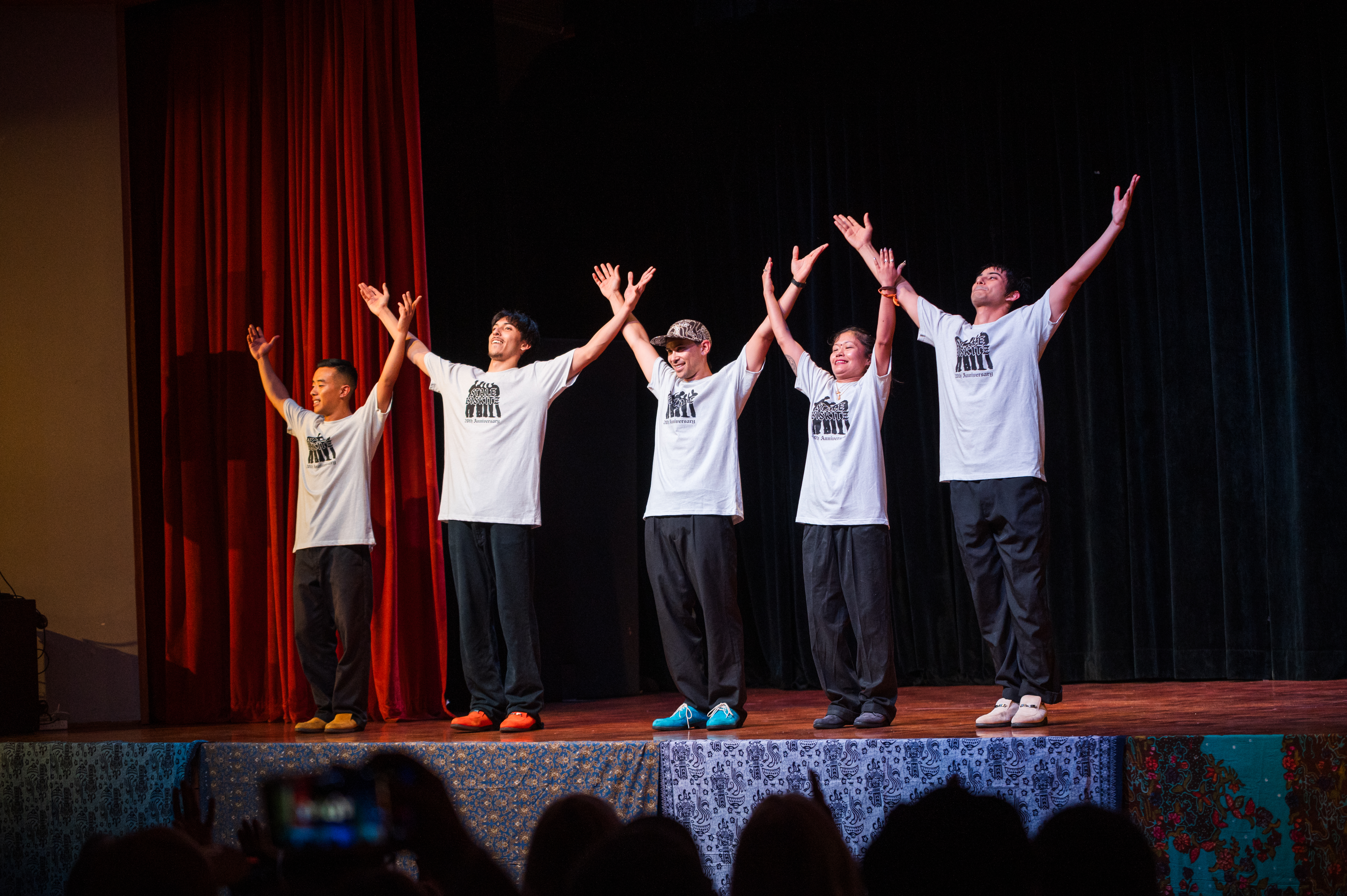 Five Stale Biskitz performers after their final performance on stage, weach smiles and has their arms raised in the air to the applause, each wear black pants and Stale Biskitz merch tshirts 