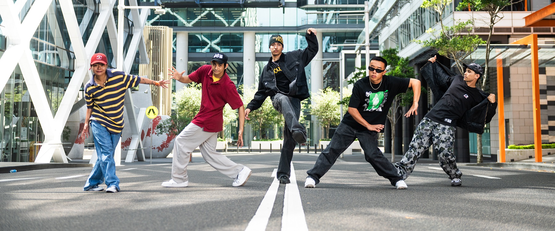 Photo of Stale Biskitz dancers posing in the street, 5 dancers pose in street dance style, they are on a road and two white lines run down the centre of the photo, corporate buildings are in the background and some trees to the side