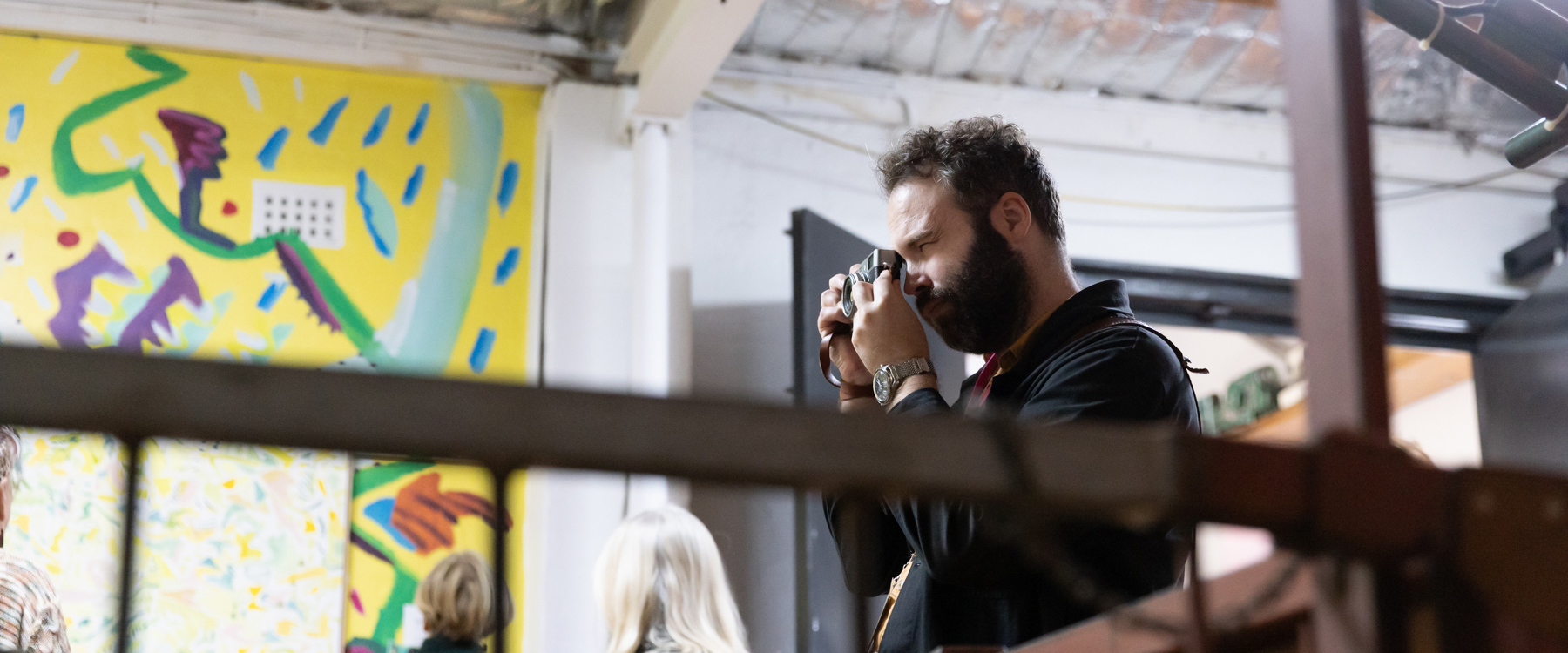 Photo of a person looking through a camera lense taking a photo, they squint and have facial hair, the background has a painting in it