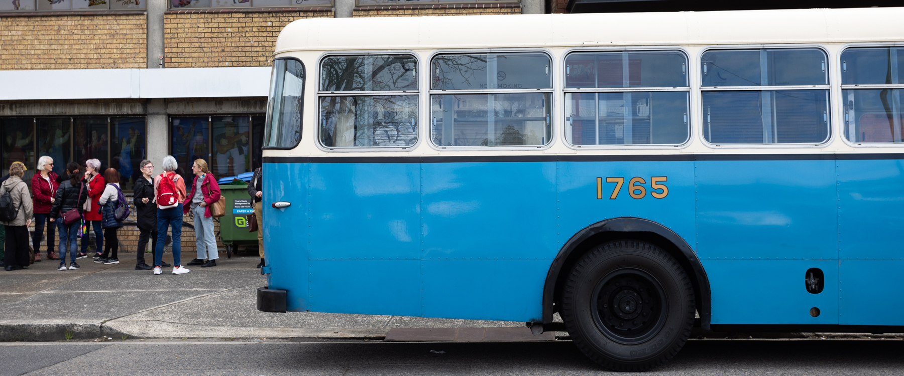 Photo of a vintage blue bus pulling out of the shot, the rear is visible and behind you can see a group of people