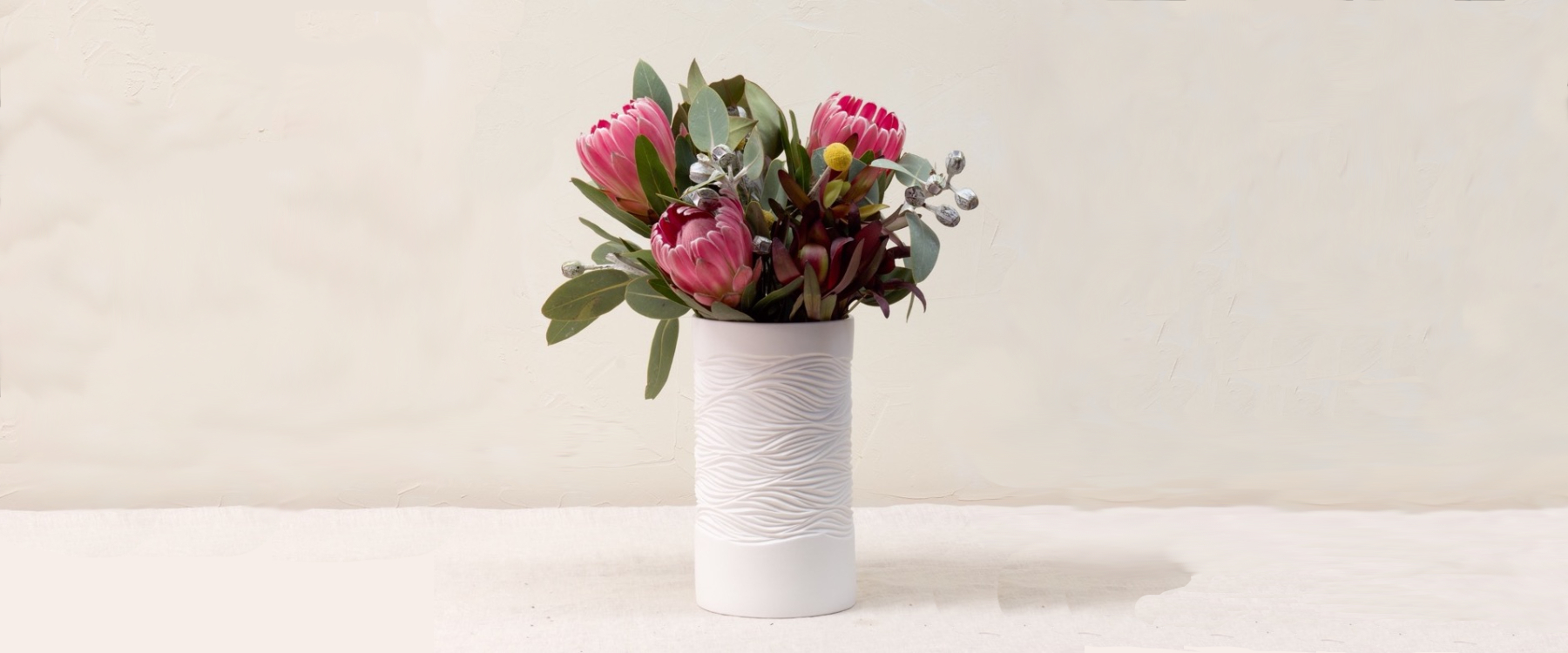 Photo of a white textured vase holding a bunch of pink Proteas, Australian native leaves, yellow button tails and silver gumnuts