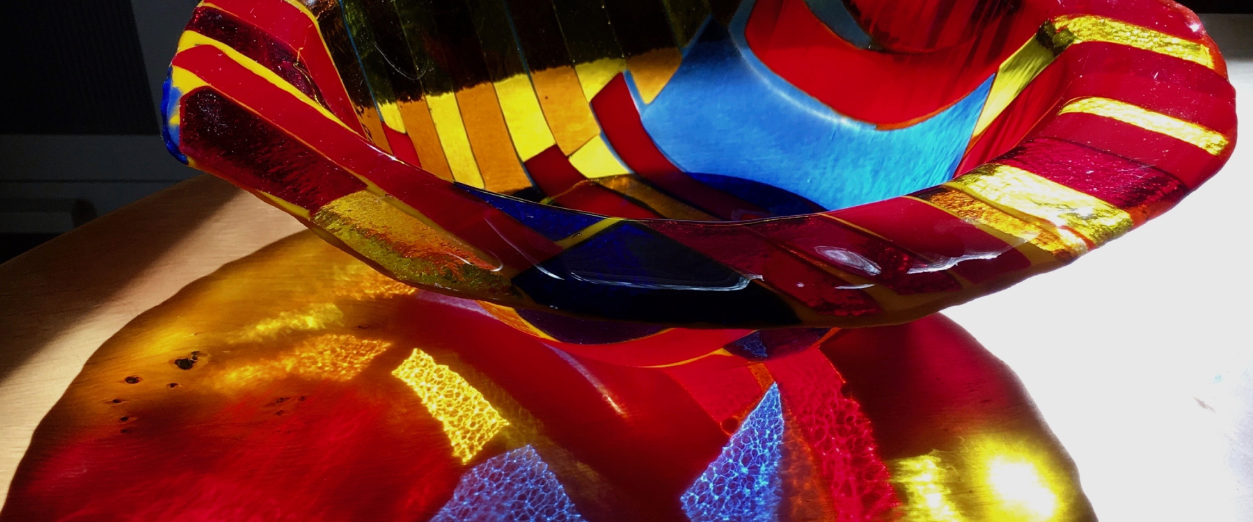 Photo of a red glass bowl, the bowl has blue and yellow shapes in it and a colourful shadow is projected onto the table it sits on