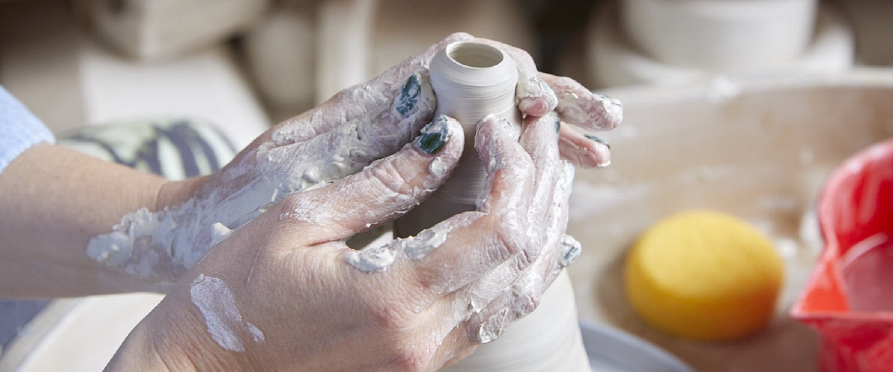 Photo of hands with green nail polish covered in clay, the hands are sculpting a vessel and it looks like it must be on a wheel, in the background is a sponge and a red bucket