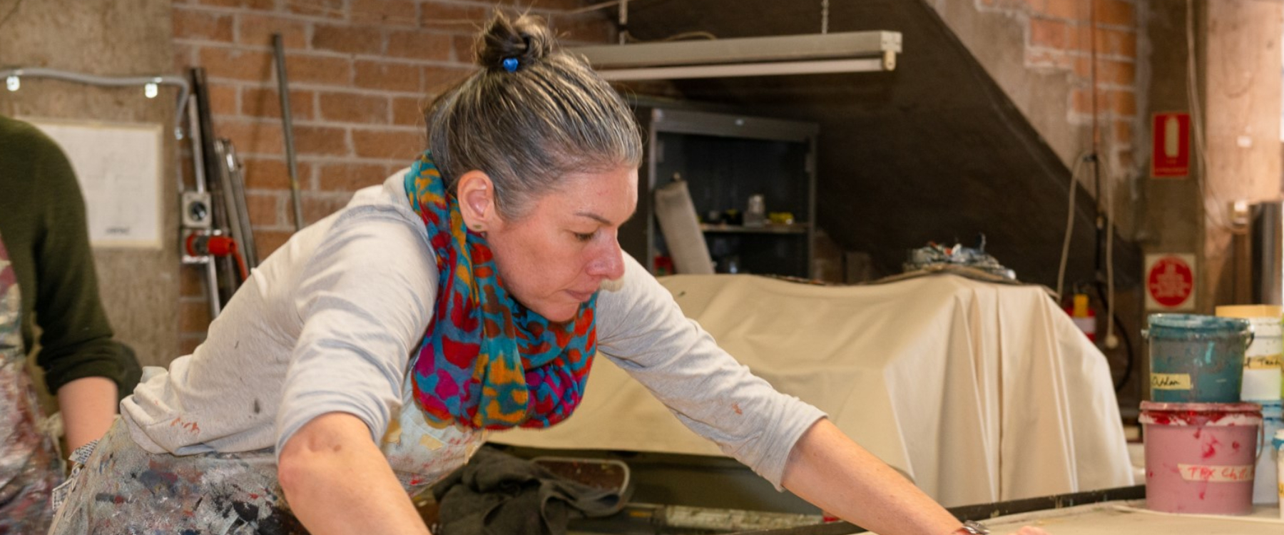 A person with a bun, apron and jumper pulls a screen print over a table, they are in the signature prints studio