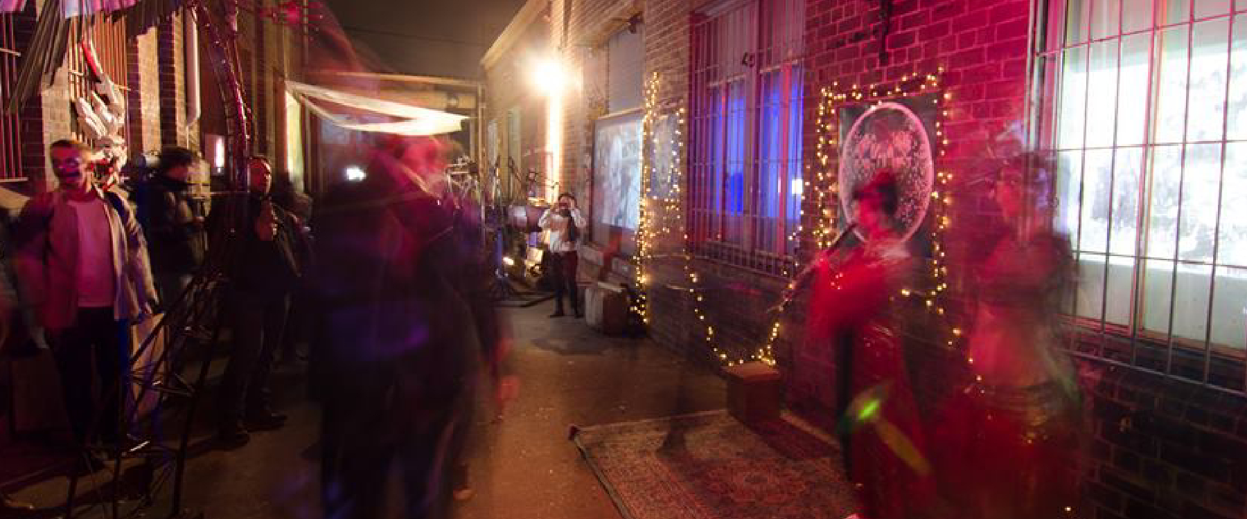 Photo of a night-time event held at the outdoor space at Tortuga Studios, there are fairy lights surrounding artworks on brick walls and some pink lighting, the photo looks like it has double exposures with movement of bodies, people are in the shot looking at artworks but not many faces are visible