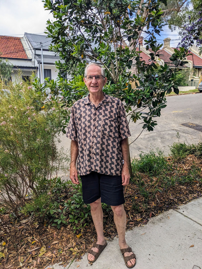 A smiling spectacled man standing in front of a verge garden.