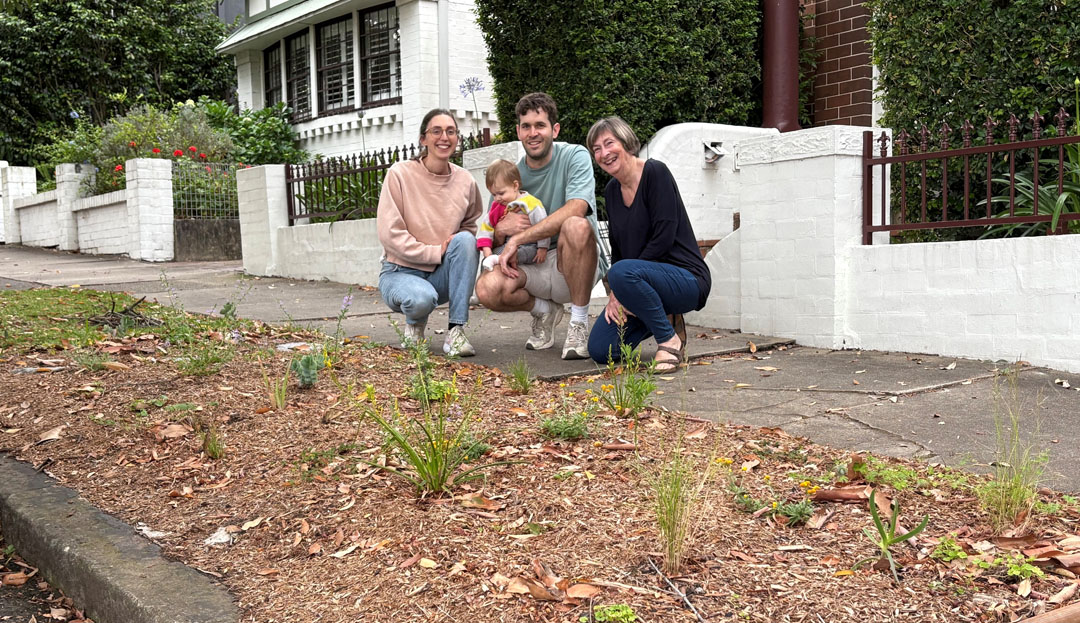 Three adults, one holding a baby, crouching next to a verge garden with a variety of young plants.