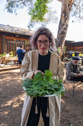 Photo of Maissa wearing an open throbe like jacket with gold and white stripes, Maissa has a large plate of fresh green outstretched grape vine leaves lying on top on each other, out of focus in the background a man sists at a table he wears a black beret and dark glasses 