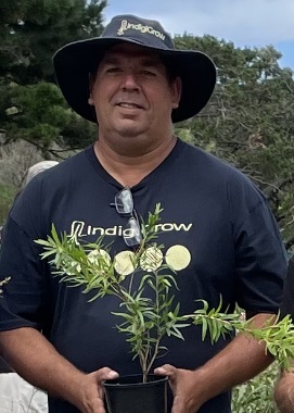 Photo of Peter Cooley outside wearing a floppy material Indigigrow hat, he holds in his hands a native plant that has ruler length fronds