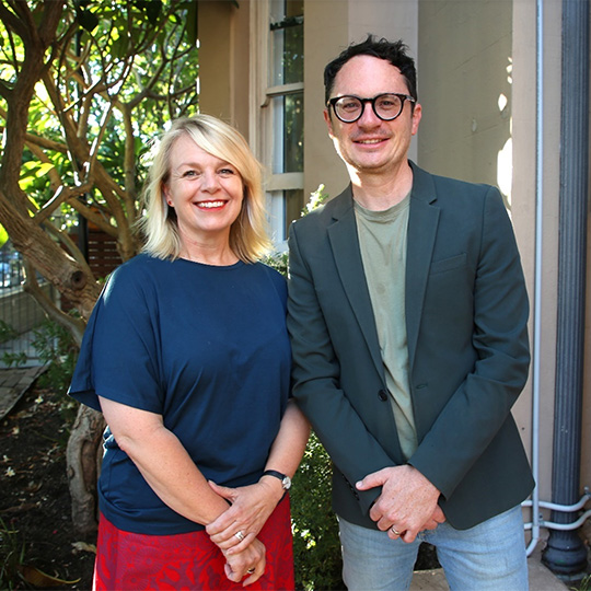 Two people smiling and standing in front of a leafy inner suburban childcare centre.