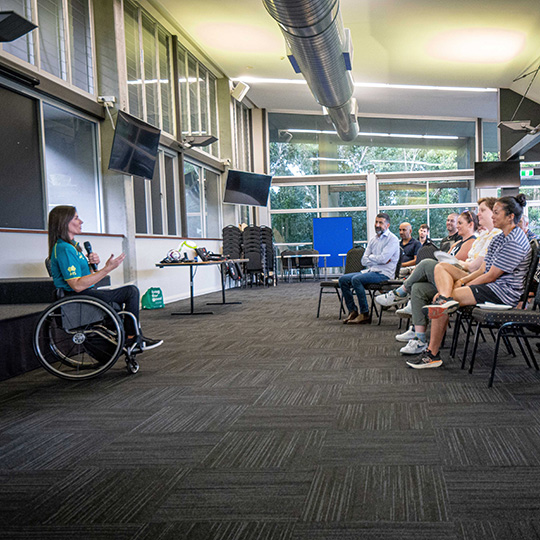 A person in a wheelchair addresses a seated audience in a function room.