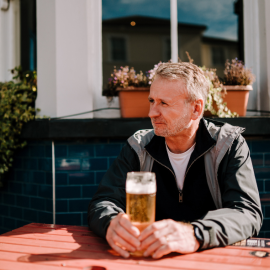 A person sits plaintively with a beer at an outdoor pub table.