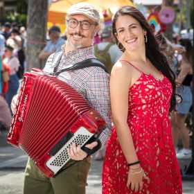 Two people smiling in the sun. One holds an accordian.