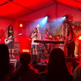 Three people perform rock songs under lights on a stage. 