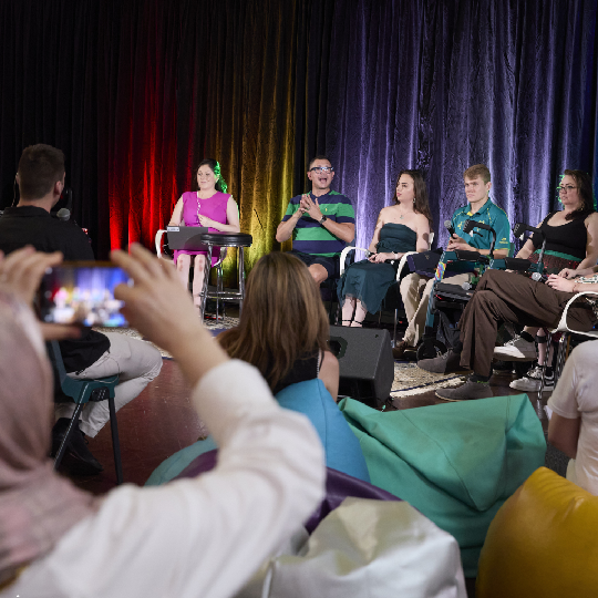 Audience watching a panel of people with disability speaking. A person in the audience wearing a headscarf is taking a photo using a mobile phone